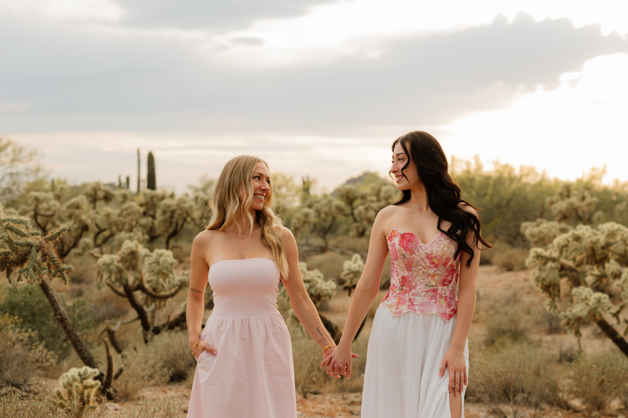 A blonde woman on the left and brunette woman on the right, holding hands looking at each other lovingly. They are mother and daughter. Standing in the desert.
