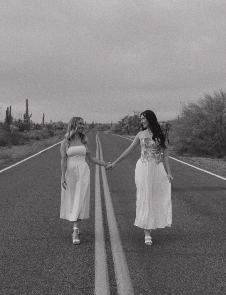 Black and white photo of a mother and daughter holding hands, looking at each other and walking in the middle of the road, in a desert.