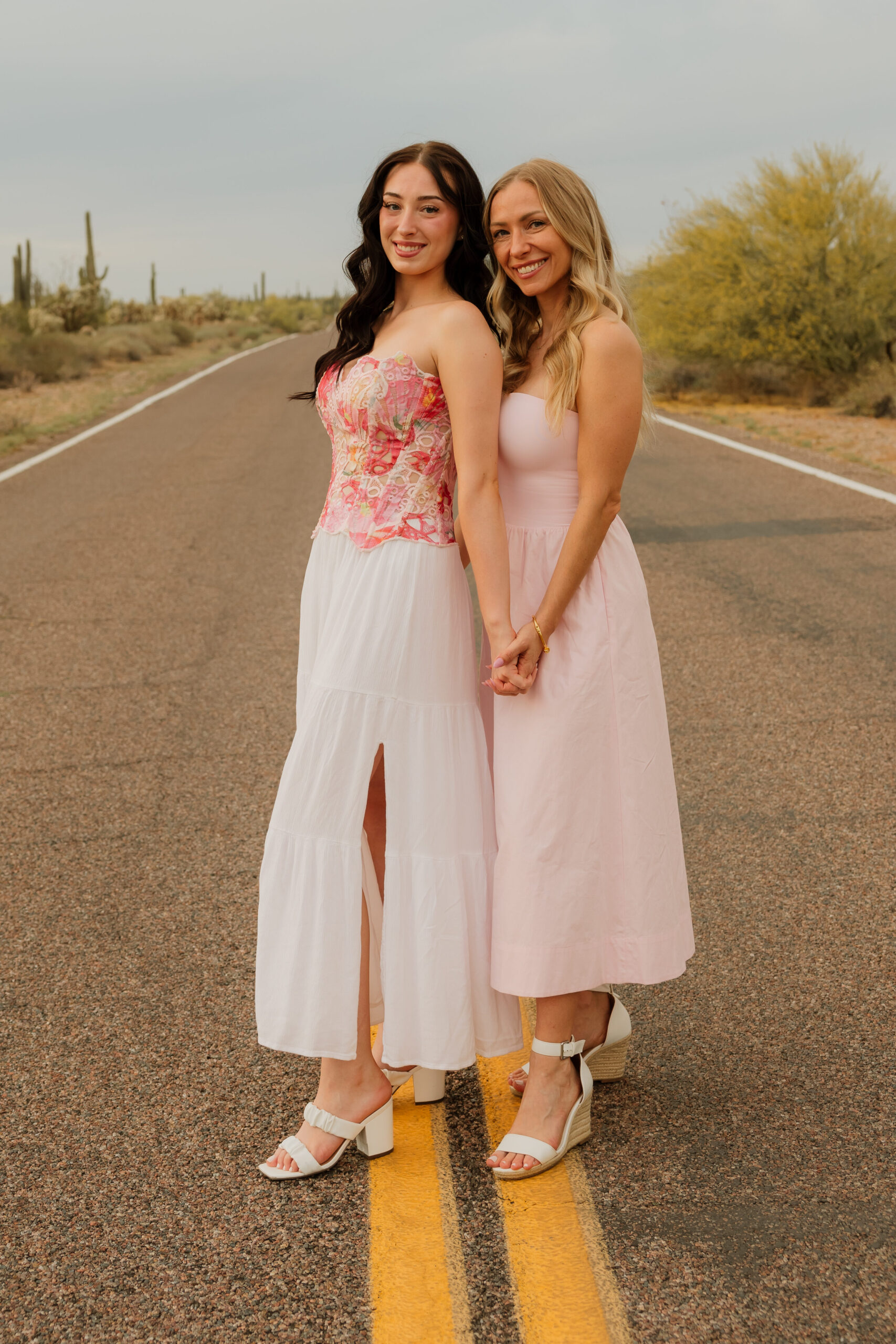 A blonde woman and a brunette woman standing together holding hands in the middle of the street, in the desert.