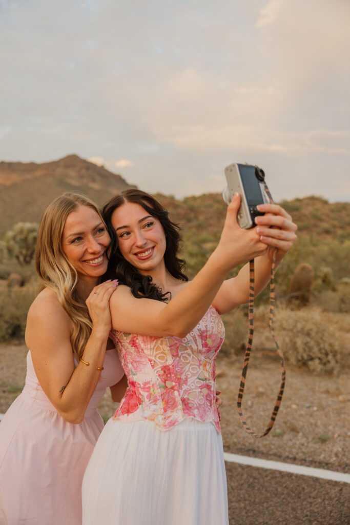 A blonde woman and a brunette woman posing for a selfie with a digital camera wearing dresses, on a road in front of a mountain. 