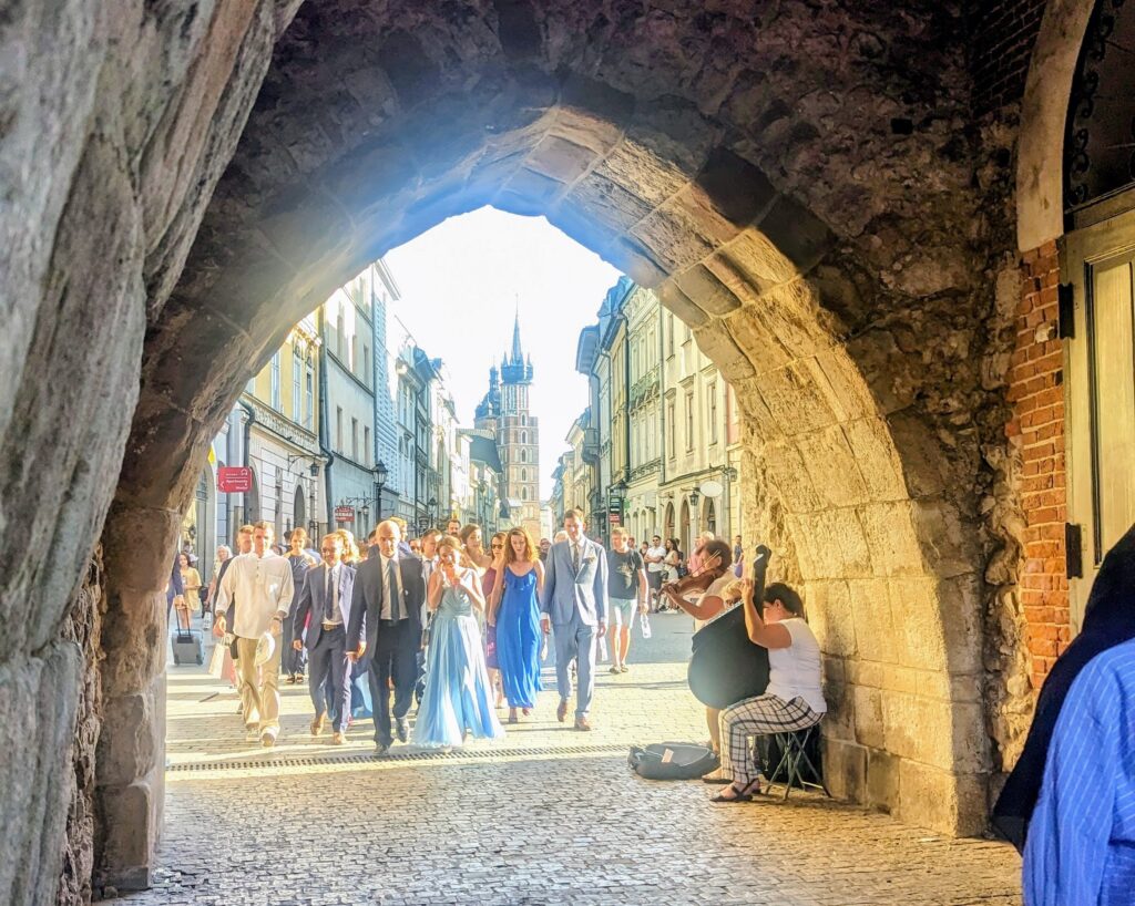 A party dressed up in formal attire walking out of Kraków main square area on a cobblestone street. St Mary's Basilica in the background, and violinists in the forefront under an archway. 