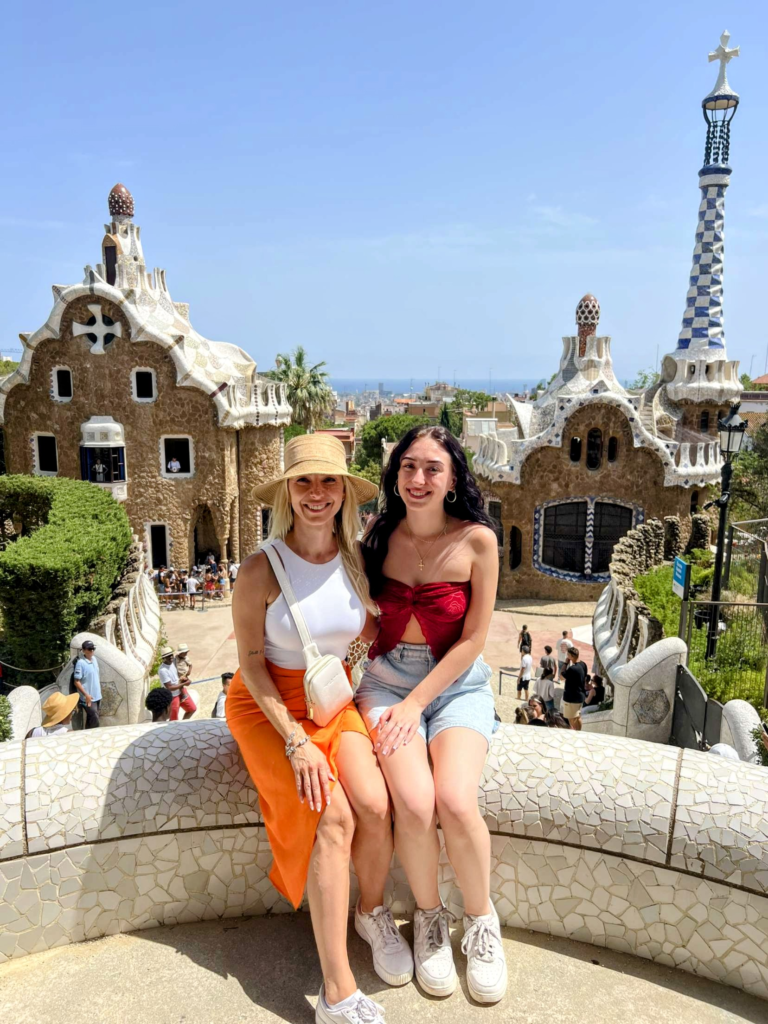 A blonde woman and brunette woman sitting on a railing at Park Güell overlooking Barcelona.