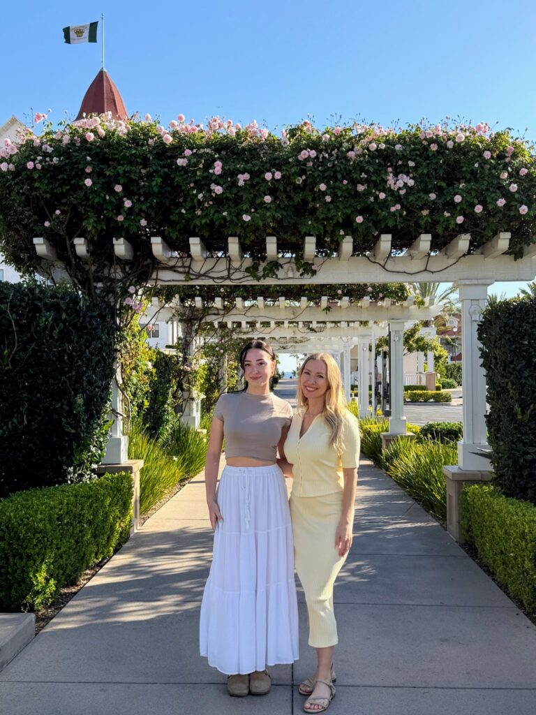 Two women standing on a sidewalk lined with pergolas draped with pink flowers. A red roof peeks over the pergola in the distance with a Corondao Island flag.