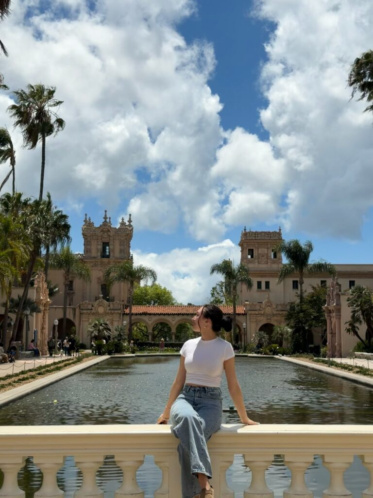 Brunette woman sitting on the railing of a bridge at Balboa Park, San Diego. The bridge is over a pond with palm trees and a building of Spanish architecture in the background.
