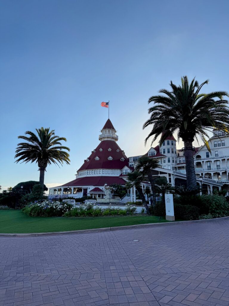 The entrance to Hotel del Coronado. A white building with a red roof and the front is a cone shaped building with the American Flag at its peak and a circular drive.